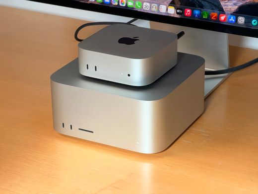 A compact silver Apple desktop stacked on a larger desktop computer beside a monitor.